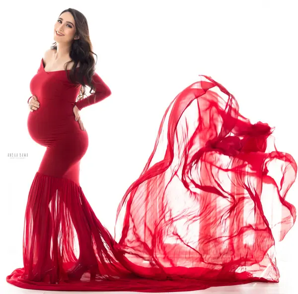 Smiling expecting mother in a red long-sleeve scoop-neck mermaid gown with a sheer ruffled hem and a sheer red train flying out to the