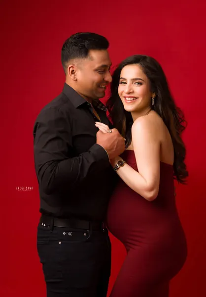 Smiling couple dancing close against a solid red backdrop, wife in a fitted red strapless gown, husband in a black shirt and jeans holding