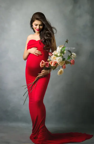 Expecting mother in a red strapless fitted gown with a long train, bright lipstick, holding a trailing bouquet of cream and peach roses