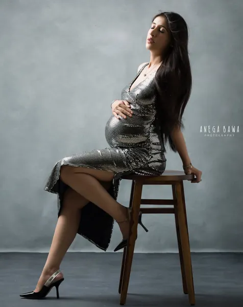 Expecting mother seated on a wooden stool in a silver metallic long-sleeve wrap dress with a slit and black pointed heels, head tilted