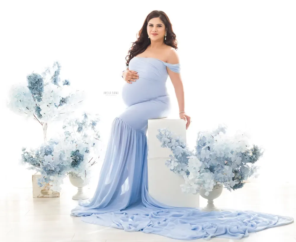 Expecting mother seated on a white block in a periwinkle blue off-shoulder mermaid gown with a long train, surrounded by pale blue