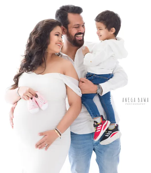 Family portrait: husband in a white shirt and jeans carrying his young son in red sneakers, wife in a white off-shoulder fitted gown