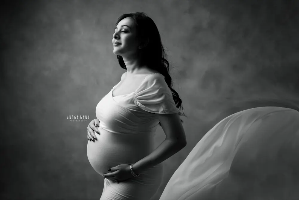 Black and white low-key portrait of expecting mother in a ruffle-sleeve fitted gown with sheer fabric at the side, looking up, hands on her