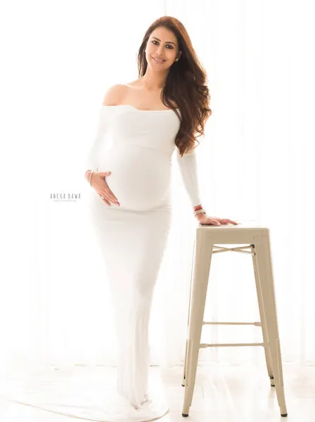 Smiling expecting mother in a white off-shoulder long-sleeve fitted gown standing beside a white metal stool, hand on her bump, bright