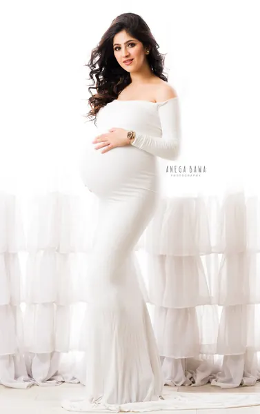 Smiling expecting mother in a white off-shoulder long-sleeve fitted gown, wavy hair, hand on her bump, in front of tiered ruffled white