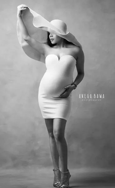 Black and white portrait of expecting mother in a white fitted strapless mini dress with a sweetheart neckline, holding up a large brimmed