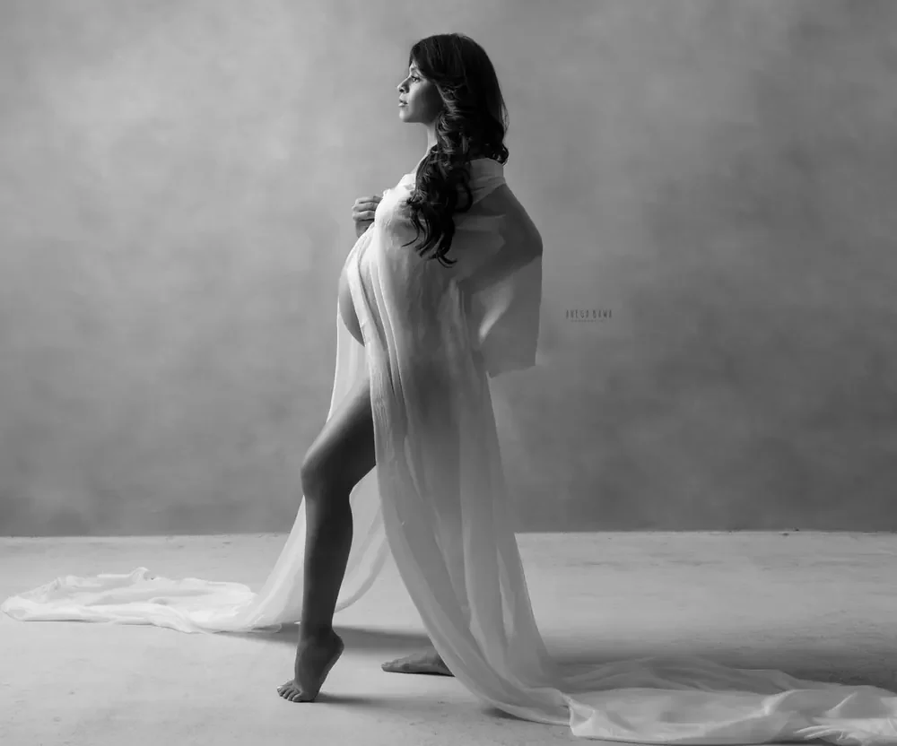 Black and white full-length profile of expecting mother in a sheer white drape with a long train on the floor, one leg forward, barefoot