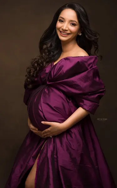 Smiling expecting mother in a plum satin gown with a gathered wrap and puffed sleeves, high slit, hands cradling her bump, dark brown