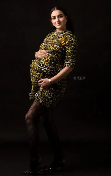 Expecting mother in a gold and black patterned knit mini dress with pearl trim, black patterned tights and heels, hand on her bump, dark