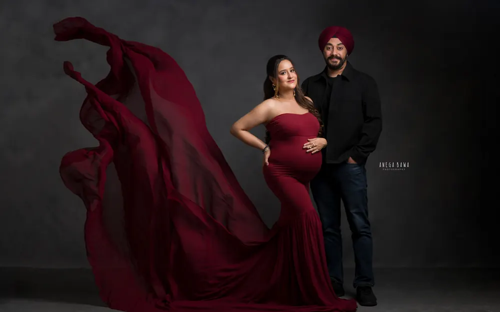 Pregnancy photography in Gurgaon: expecting mother in a maroon strapless mermaid gown with a long sheer train flowing behind her, husband