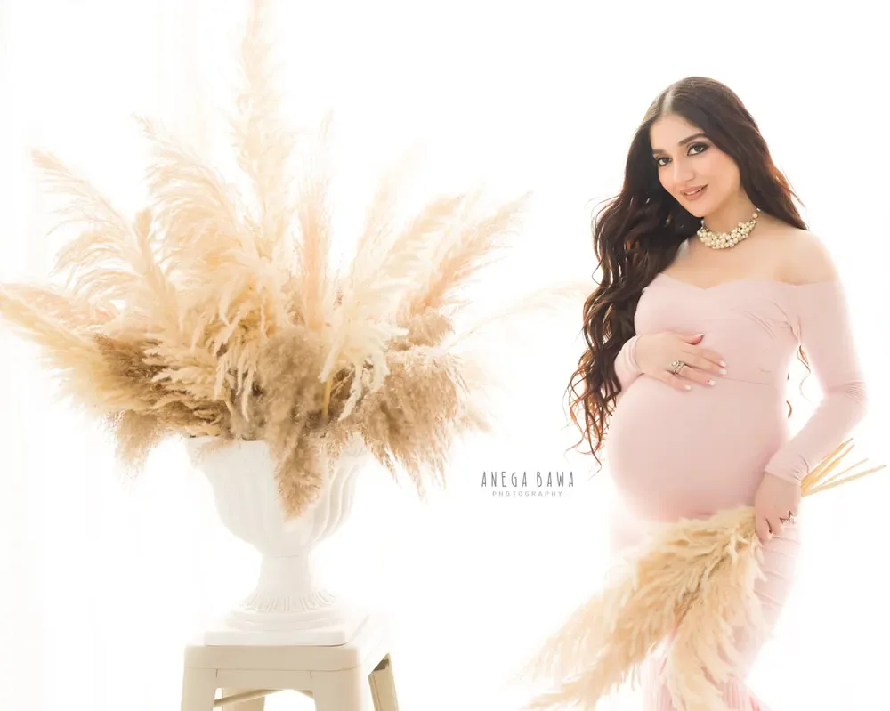 Expecting mother in a blush pink off-shoulder long-sleeve fitted dress with a gold necklace beside a white urn of pampas grass, holding
