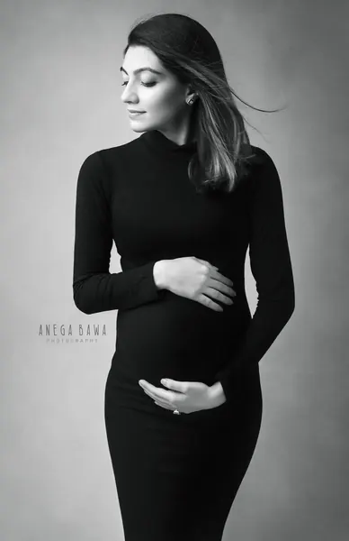 Black and white portrait of expecting mother in a black turtleneck bodycon dress, eyes down, hair blowing, hands cradling her bump, light