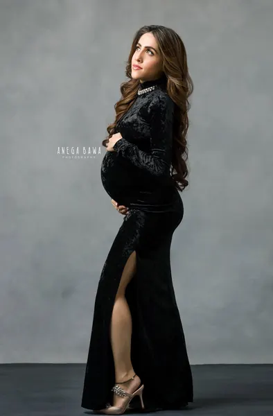 Expecting mother in profile in a black velvet high-neck long-sleeve fitted gown with a pearl collar and thigh slit and embellished heels