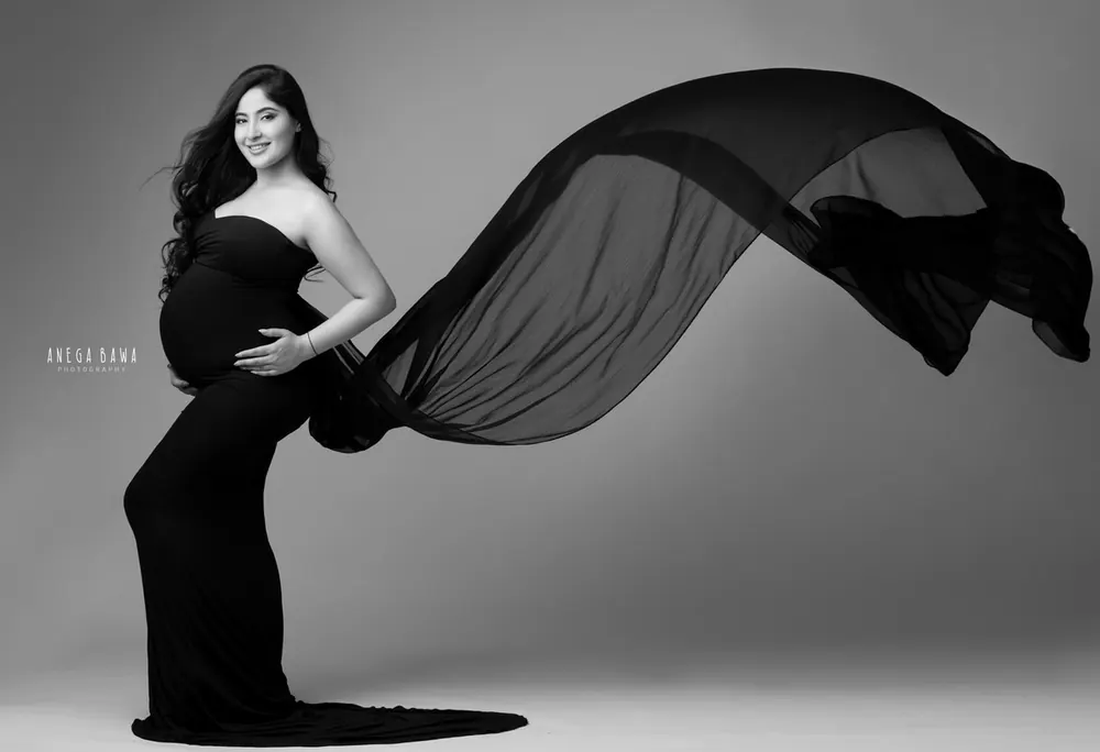 Black and white portrait of expecting mother in a fitted black strapless gown with a long sheer black train flying out to one side, hand on