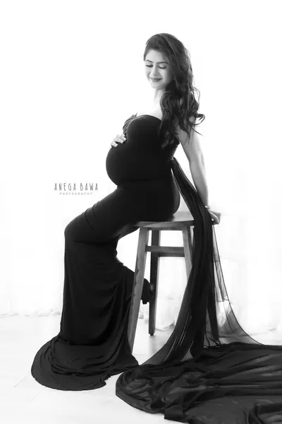 High-key black and white portrait of expecting mother seated on a wooden stool in a black strapless gown with a long sheer train, looking