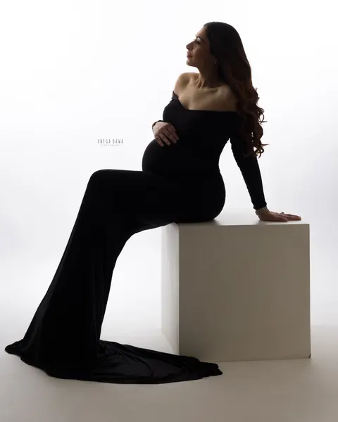 Pregnancy photoshoot in Delhi: backlit silhouette of expecting mother seated on a white block in an off-shoulder black gown with a long