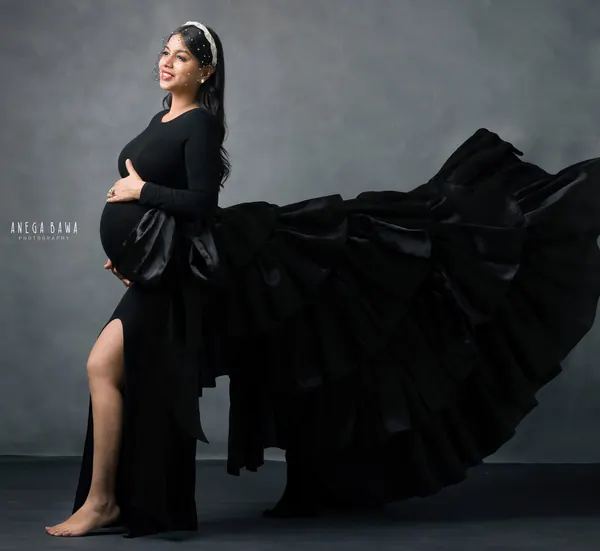 Expecting mother in a black long-sleeve fitted dress with a high slit and a huge tiered black ruffle train swept out behind her, headband