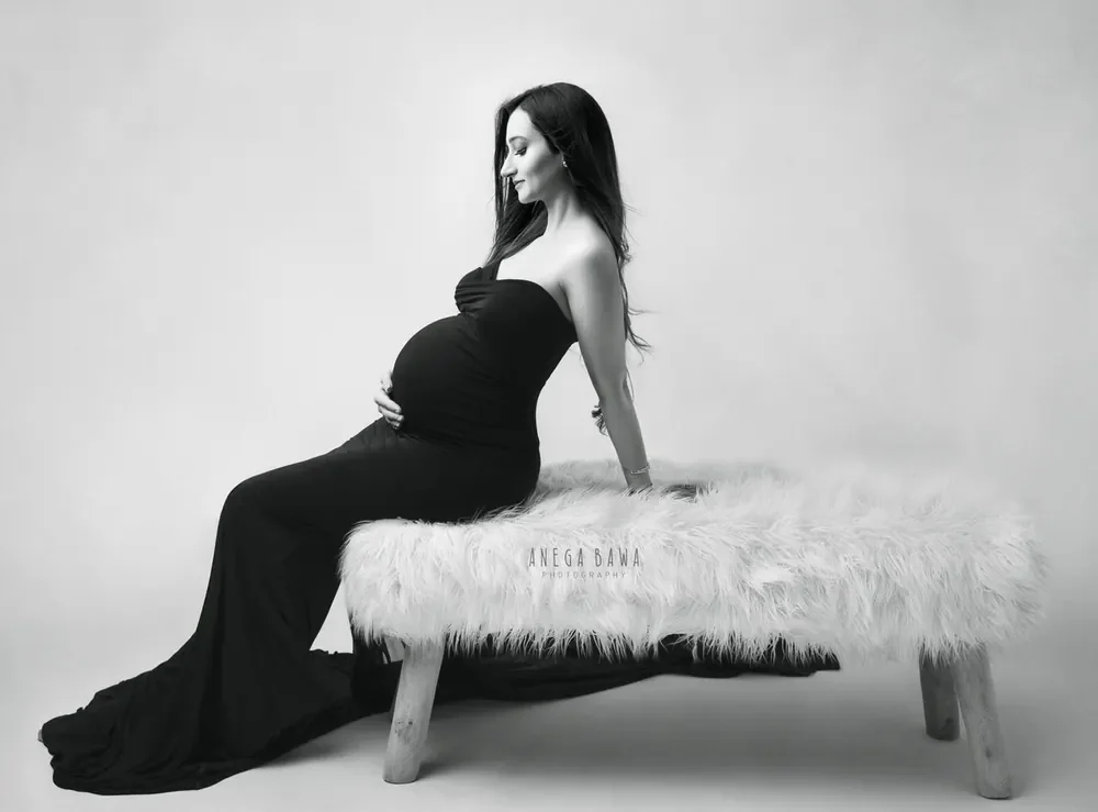 Black and white portrait of expecting mother seated on a white fur bench in a fitted black strapless gown with a long train, looking down