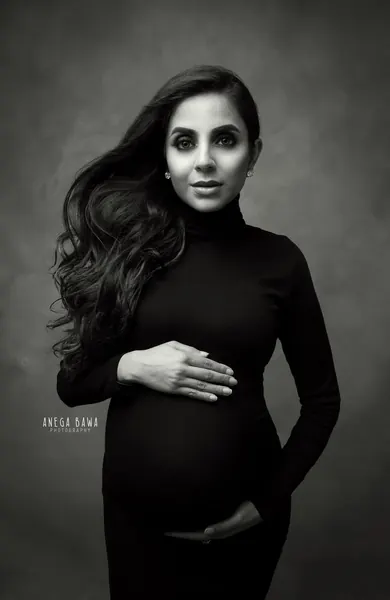 Black and white portrait of expecting mother in a black turtleneck fitted dress with voluminous wavy hair, one hand on her bump and one