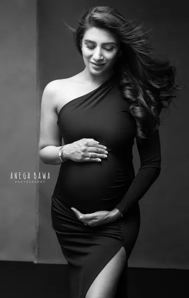 Black and white portrait of expecting mother in a black one-shoulder long-sleeve ruched gown with a high slit, hair blowing, eyes down