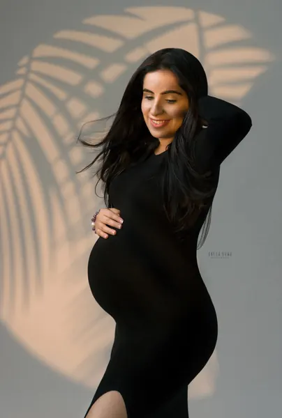 Smiling expecting mother in a fitted black long-sleeve dress with a side slit, one hand behind her head and one on her bump, long hair