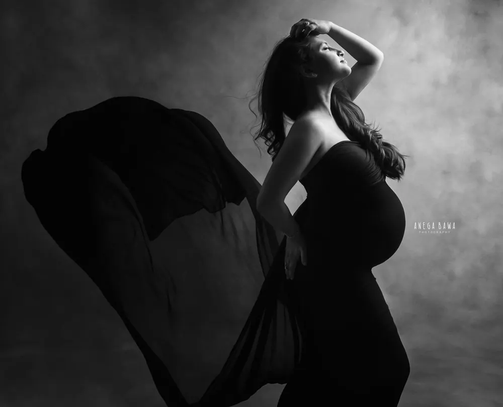 Black and white portrait of expecting mother in a strapless black gown with a sheer black drape billowing beside her, one hand in her hair