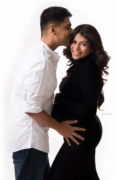 Smiling couple, husband in a white shirt kissing his wife's head and holding her bump, wife in a black turtleneck fitted dress, bright
