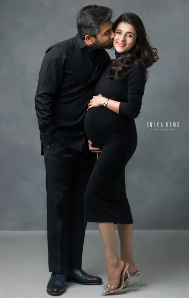 Couple portrait: husband in a black shirt and trousers kissing his smiling wife's cheek, wife in a black long-sleeve midi dress and clear