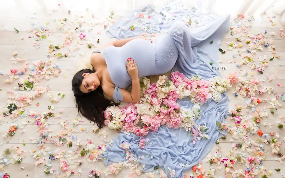 Pregnancy photoshoot in Delhi: overhead view of expecting mother lying on a wooden floor in a powder blue gown surrounded by pink peonies