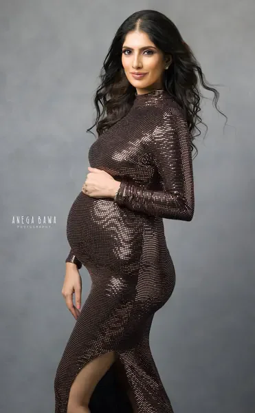 Expecting mother in a bronze sequin high-neck long-sleeve fitted gown with a thigh slit, long wavy hair, hand on her bump, looking at the