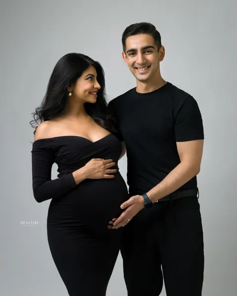 Pregnancy photoshoot in Delhi: smiling couple in all-black outfits, wife in an off-shoulder gown looking at her husband whose hand rests on