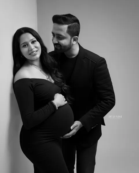 Pregnancy photoshoot in Delhi: black and white portrait of a couple in black, wife leaning against a studio wall in an off-shoulder gown