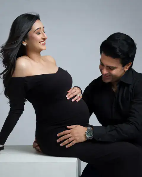 Pregnancy photoshoot in Delhi: laughing wife seated on a white block in a black off-shoulder gown while her husband in black leans in with