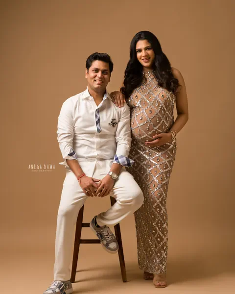 Pregnancy photoshoot in Delhi: couple posing together, wife in a crystal-embellished halter gown and husband in white seated on a wooden
