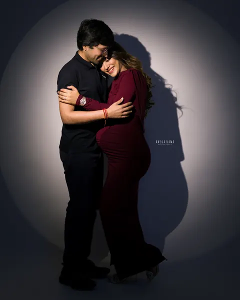 Pregnancy photoshoot in Delhi: couple embracing in a circle of spotlight, wife in a burgundy gown and husband in a black polo, by Anega