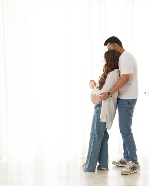 Pregnancy photoshoot in Delhi: husband in a white tee and jeans hugging his wife from behind, both looking down at her bump, sheer white