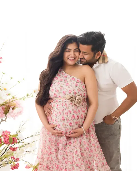 Pregnancy photoshoot in Delhi: laughing wife in a floral-print dress with her husband in a white polo kissing her cheek, hands on her bump