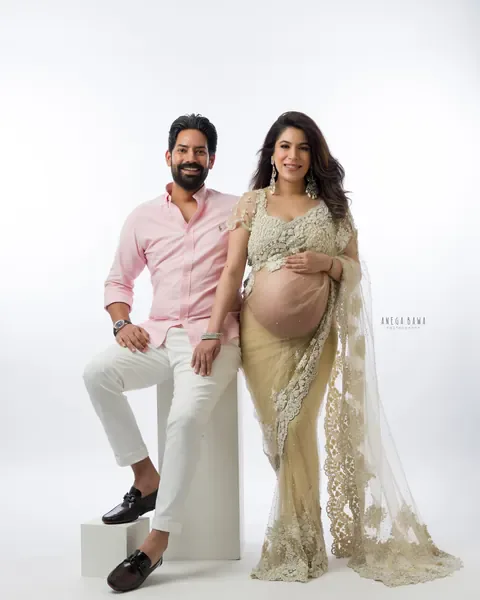 Pregnancy photoshoot in Delhi: smiling couple, husband in a pink shirt and white trousers seated on a white block beside his wife in a