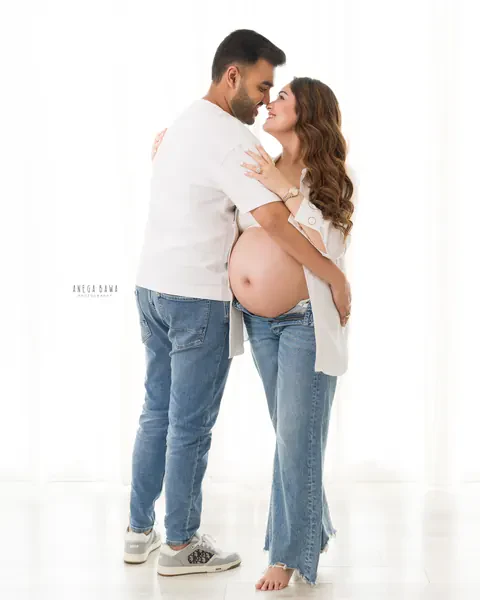 Pregnancy photoshoot in Delhi: couple standing nose to nose, husband in a white tee and wife in an open white shirt and jeans with her bare