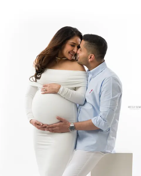 Pregnancy photoshoot in Delhi: husband in a light blue shirt nuzzling his smiling wife who wears a fitted white off-shoulder gown, hands on