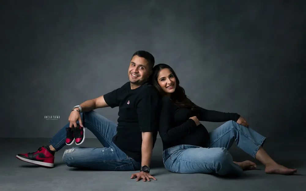 Pregnancy photoshoot in Delhi: couple seated back to back on the studio floor in black tops and jeans, husband holding tiny baby sneakers