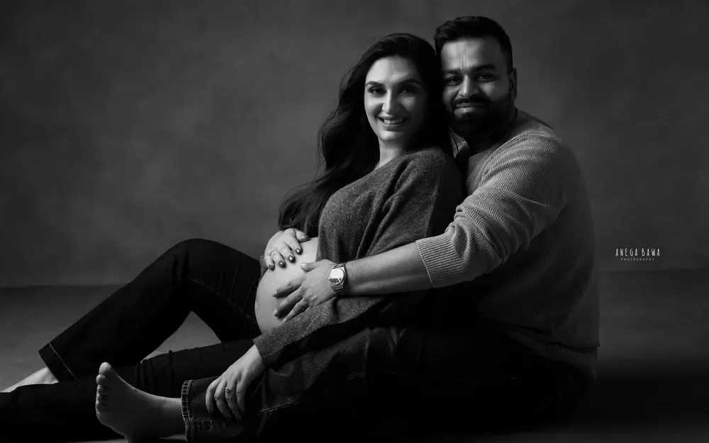 Pregnancy photoshoot in Delhi: black and white portrait of a smiling couple seated on a studio floor, husband's hands on her bump, by Anega