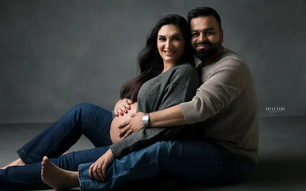 Pregnancy photoshoot in Delhi: smiling couple seated on a studio floor in jeans and grey knit tops, husband's hands on her bump, by Anega