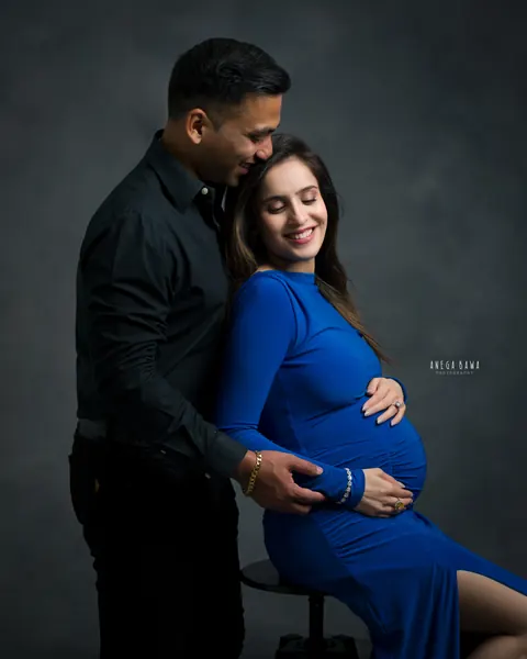 Pregnancy photoshoot in Delhi: husband in a black shirt kissing his wife's hair as she sits on a stool in a royal blue gown, both hands on