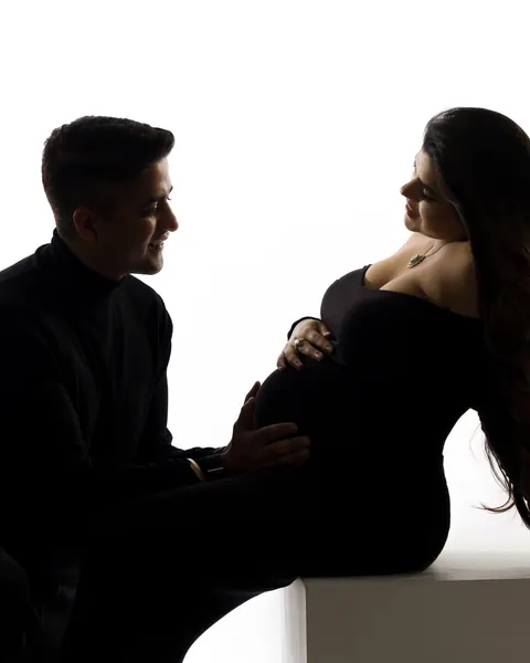 Pregnancy photoshoot in Delhi: backlit silhouette of a husband kneeling to touch his wife's bump as she sits on a white block in a black