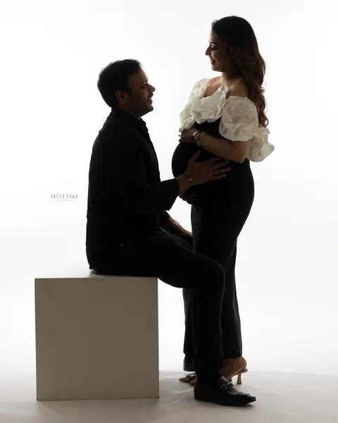 Pregnancy photoshoot in Delhi: backlit couple facing each other, husband seated on a white block and wife standing in a black outfit with