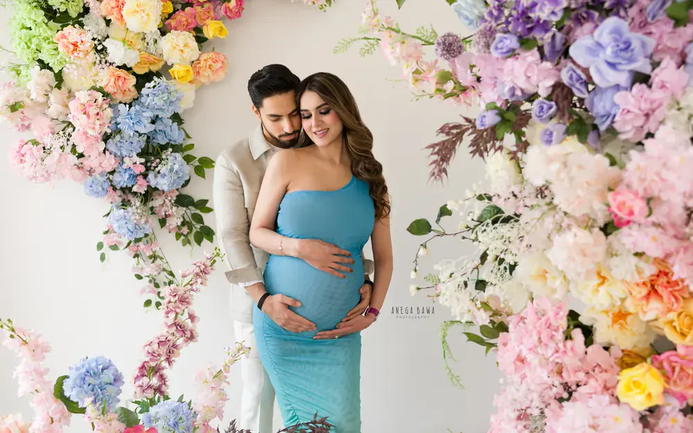 Pregnancy photoshoot in Delhi: couple embracing under a pastel flower arch, wife in a teal ruched gown and husband in a linen jacket, hands