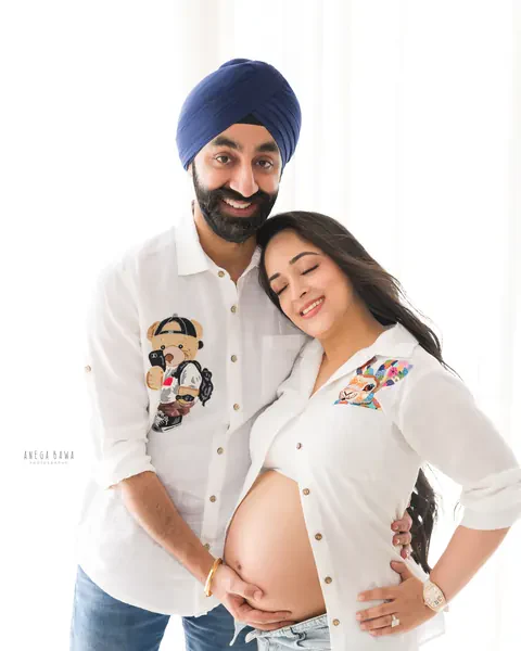 Pregnancy photoshoot in Delhi: smiling couple in white embroidered shirts, husband in a navy turban with his hand on her bare bump, white