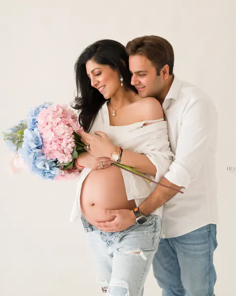 Pregnancy photoshoot in Delhi: couple in white shirts and light denim holding a bouquet of pink and blue hydrangeas, husband hugging her