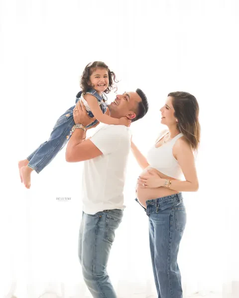 Pregnancy photoshoot in Delhi: father lifting his laughing toddler in the air while the expecting mother looks on with a hand on her bare